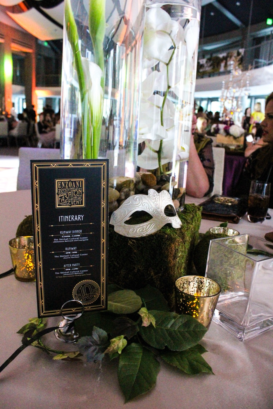 2015 Enzoani itinerary card beside a masquerade mask, orchids, and gold votives on a dinner table